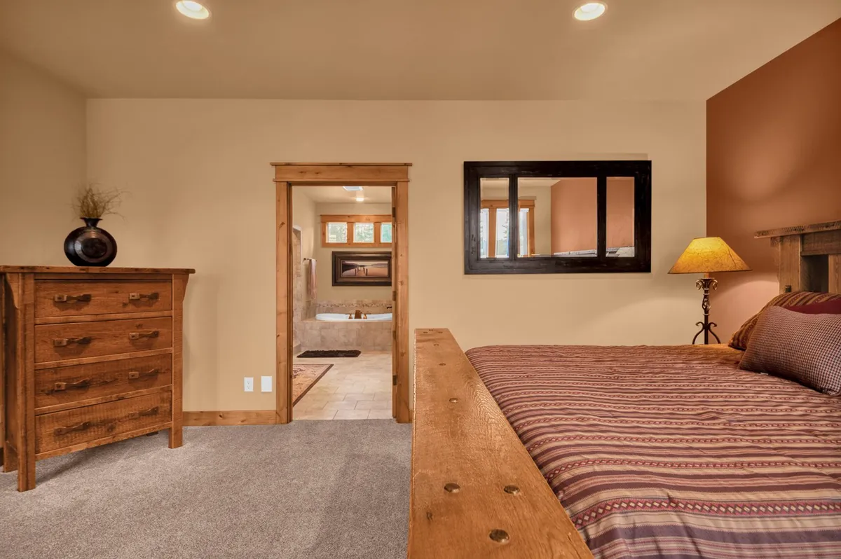 A restful bedroom with a striped bedspread, a wooden dresser, a bedside lamp, and a view of an adjacent bathroom through an open doorway. The space feels calm, intimate, and perfectly designed for peaceful nights or quiet mornings.