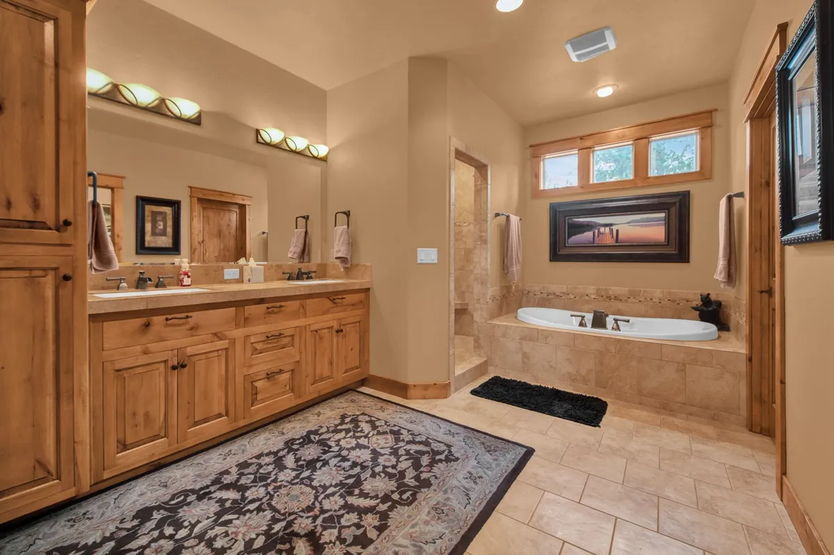 A serene, spa-like bathroom featuring a double vanity with wooden cabinetry, a luxurious freestanding bathtub, a large walk-in shower, an intricate decorative rug, and framed artwork on the wall. The space is airy and inviting, perfect for relaxation and self-care routines.