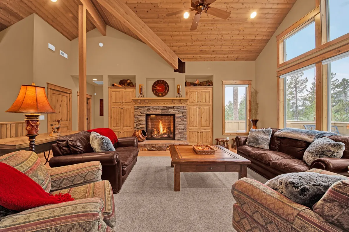 A spacious, sunlit living room featuring a vaulted wood-clad ceiling, comfortable sofas arranged around a striking stone-faced gas fireplace, and large windows that flood the area with natural light. The open flow to the adjacent spaces hints at connectivity with the dining and kitchen areas.
