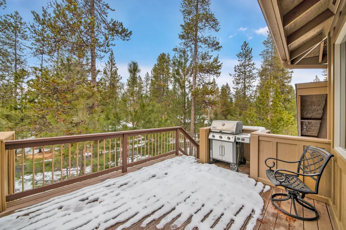 A spacious deck with a grill, surrounded by snow-covered flooring and framed by towering trees, with a scenic mountain view in the background. The area is set up for outdoor dining and socializing, offering a tranquil spot for relaxation and gatherings.