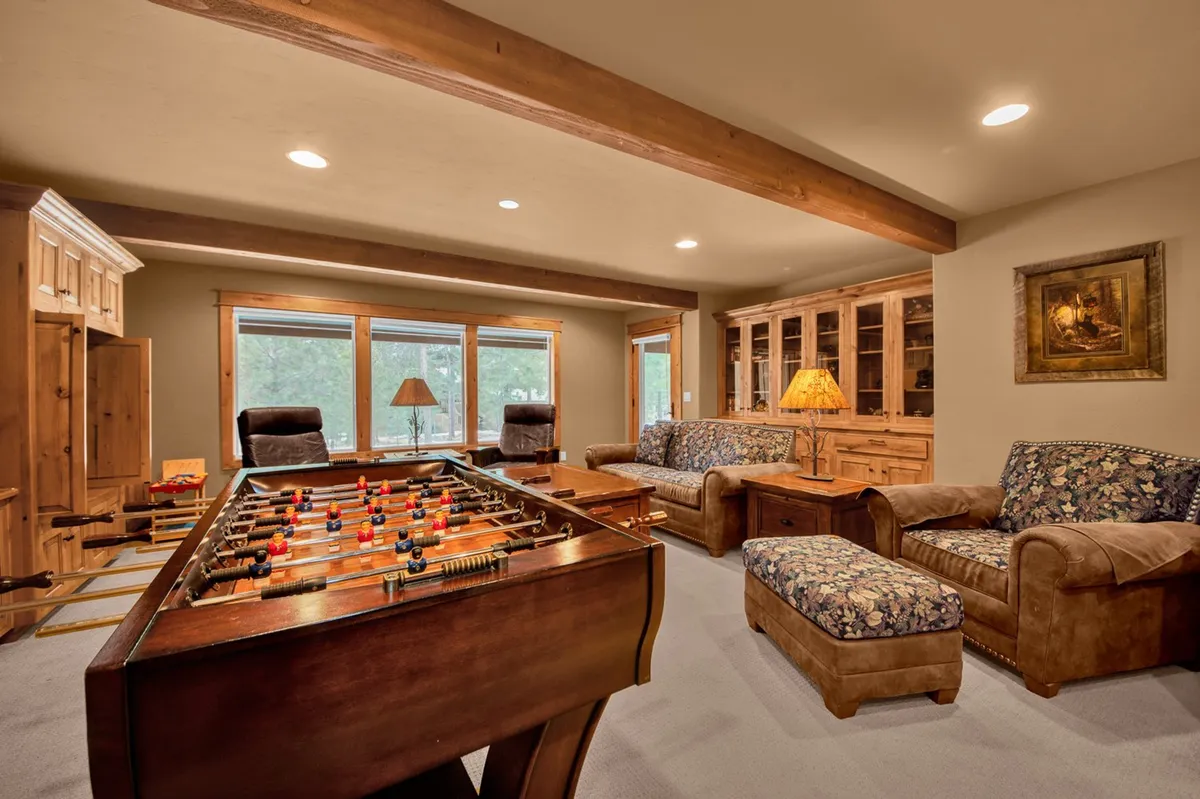 A lively downstairs living room featuring a foosball table positioned near a set of large windows, a comfortable sofa set, and rustic wooden furniture. The abundance of natural light and open space suggests a perfect environment for games, relaxation, and family interaction.