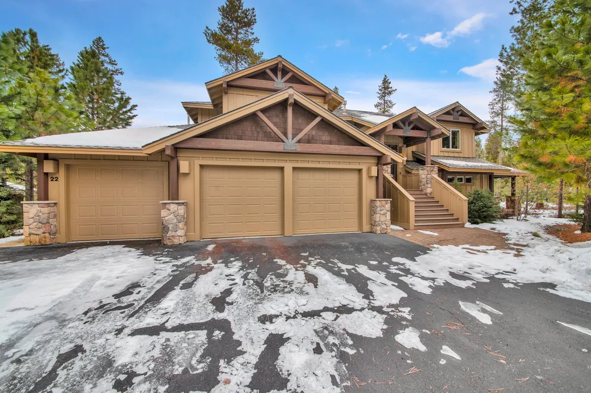 A welcoming exterior view of the home's three-car garage and main entrance, framed by tall trees and featuring wide stairs leading up to the wooden architecture. The surrounding landscape hints at space for outdoor gatherings and play, making it inviting for families with children.