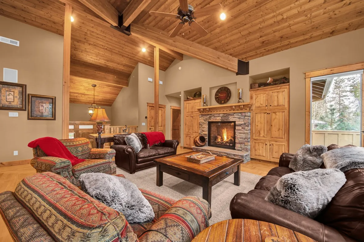 The image depicts a spacious, warmly-lit living area with exposed wooden beams, a glowing fireplace, and plush, inviting seating. The setting exudes comfort and encourages gatherings, relaxation, and intimate moments, reflecting the heart of a welcoming family vacation home.