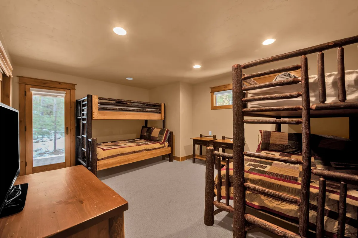 A cozy bunk room with two log-style bunk beds, warm wooden furniture, a TV for entertainment, and ample windows letting in light. The arrangement creates a camp-like, inviting atmosphere ideal for kids or groups.