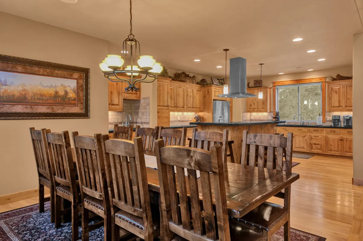 A large wooden dining table sits beneath a glowing chandelier, surrounded by kitchen cabinetry and a window that offers exterior views. The space is set up for communal meals, socializing, and sharing stories after exploring Sunriver.