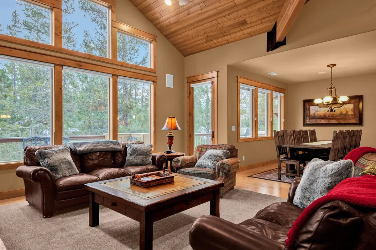 A spacious living area with leather sofas arranged around a wooden coffee table, large windows that provide ample natural light and views outside, and a dining area visible in the background. The setting feels warm and inviting, suggesting a hub for relaxing or gathering after a day of adventure.