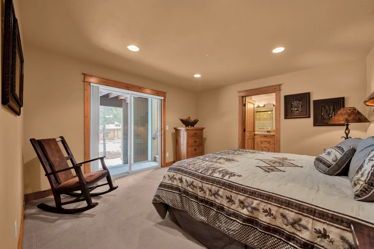 This image captures a tranquil bedroom featuring a bed with decorative bedding, a wooden rocking chair, a sliding glass door leading directly to an outdoor area, and a wooden dresser. The presence of natural light streaming inside and the seamless connection to the outdoors suggest easy access to the scenic surroundings of Sunriver.
